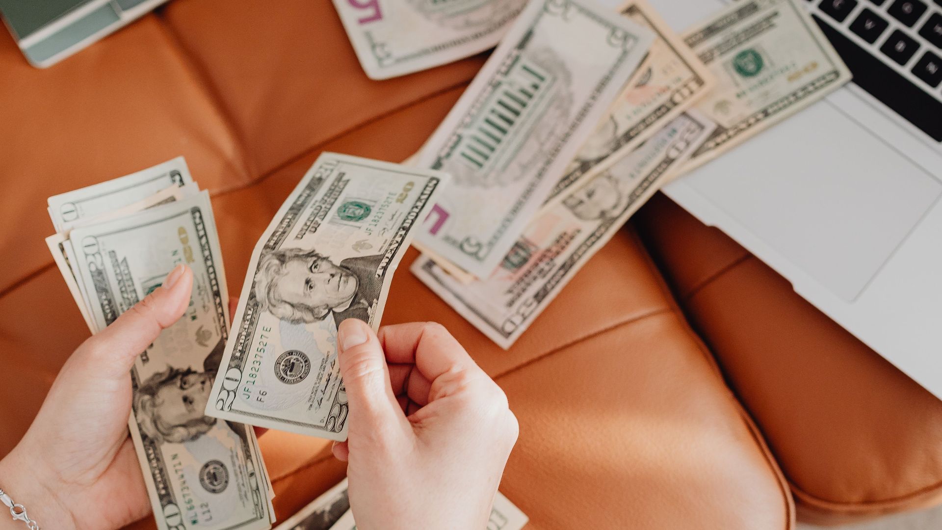 Person counting cash with a laptop and notepad on leather couch, representing finance and budgeting.
