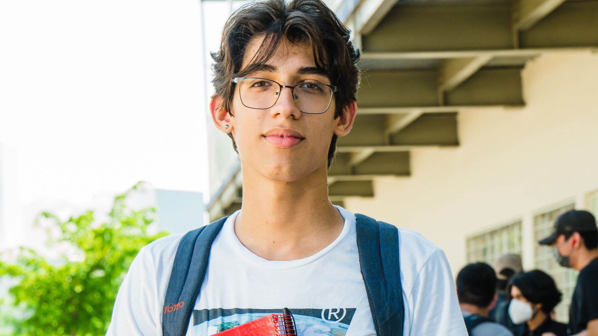 Hispanic university student outdoors in Culiacán holding a red notebook, wearing casual attire.