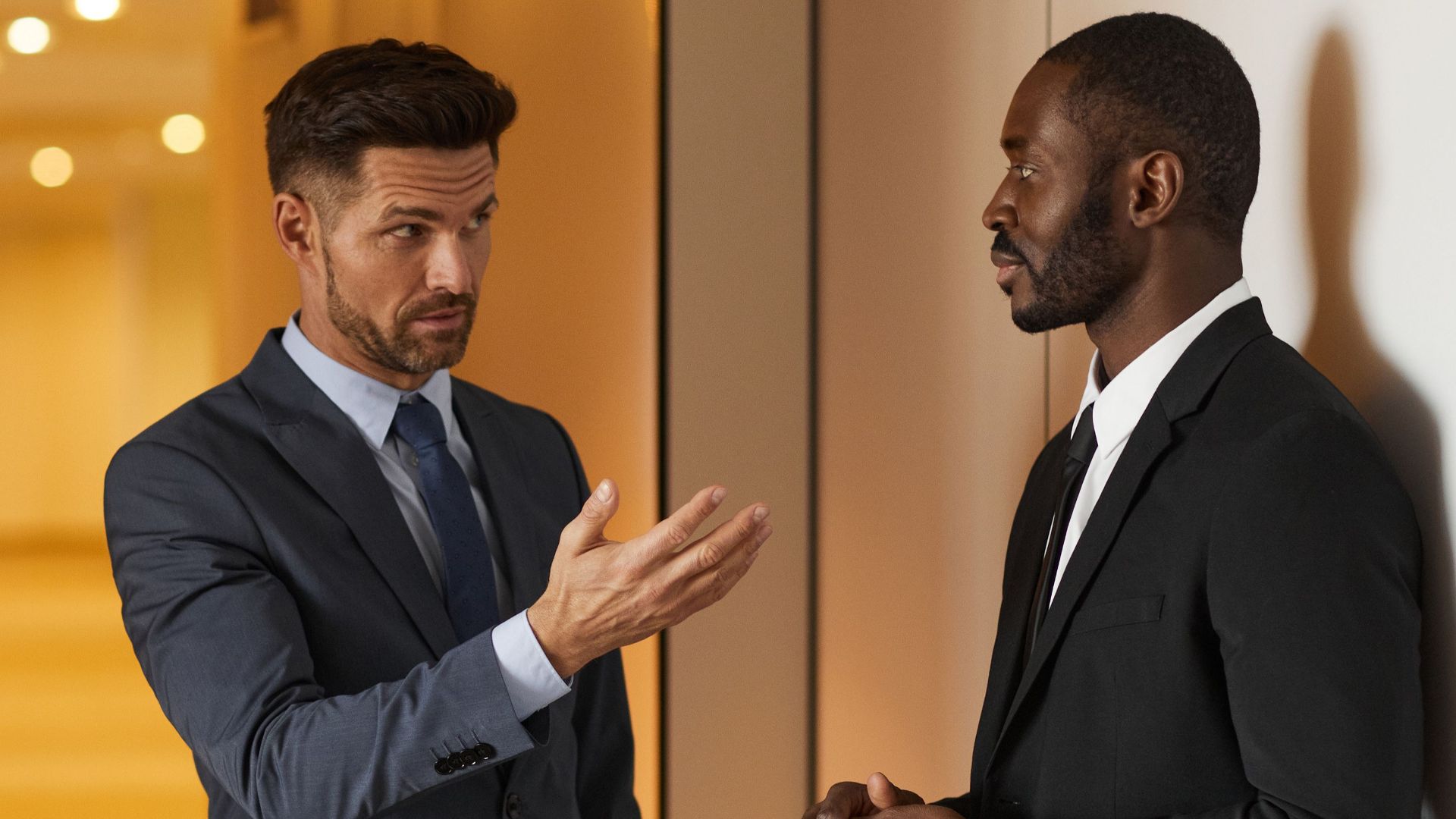 Two businessmen in formal attire engaged in a serious discussion in a modern office hallway.