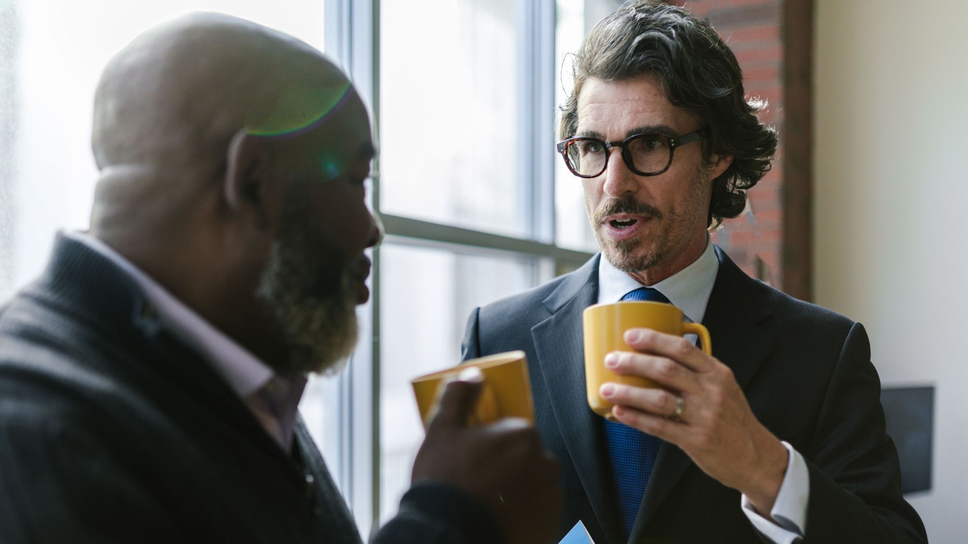 Two diverse professionals in corporate attire having a casual conversation over coffee indoors.