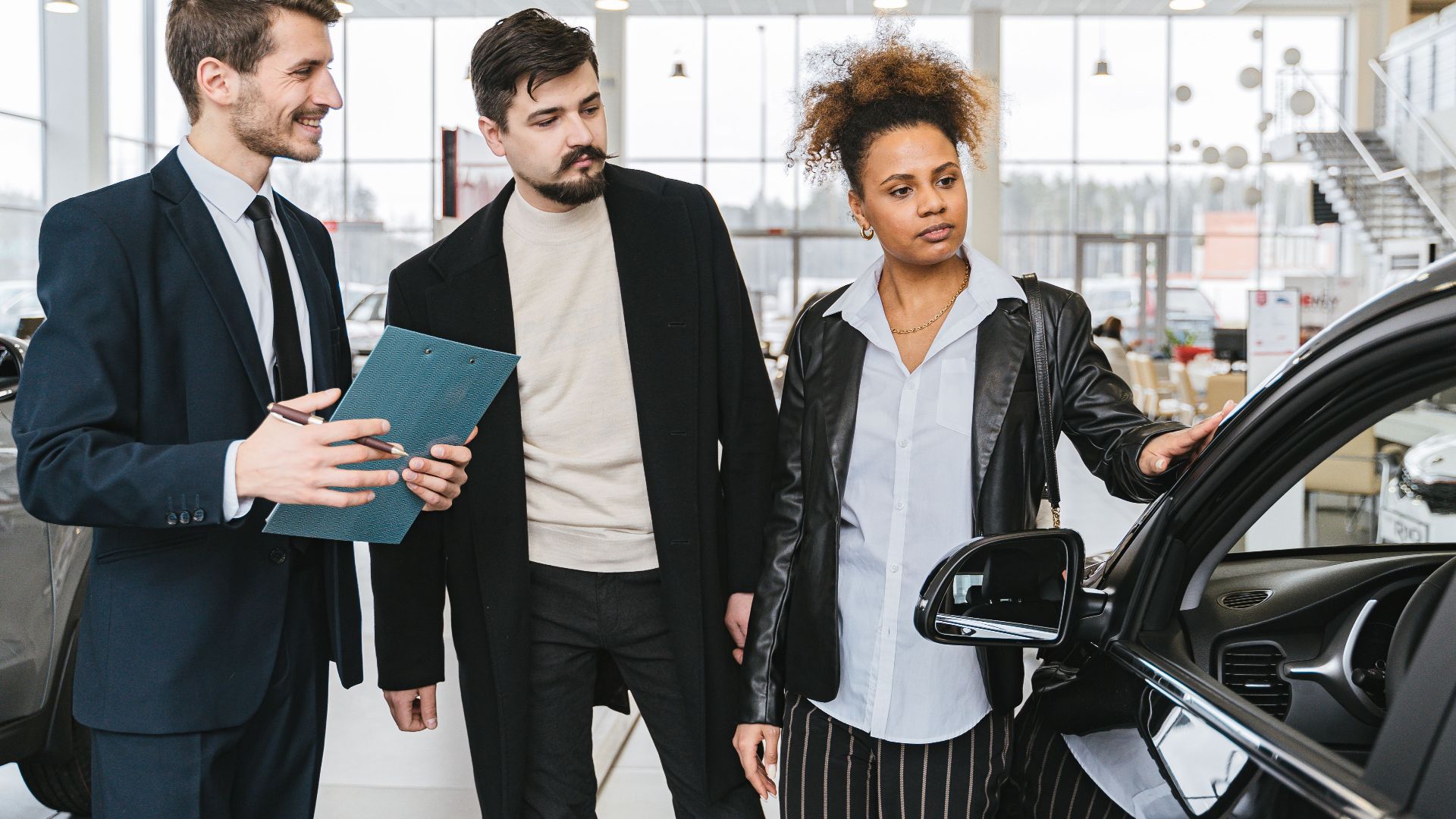 An interracial couple consults with a salesman at a car dealership, exploring vehicle options.