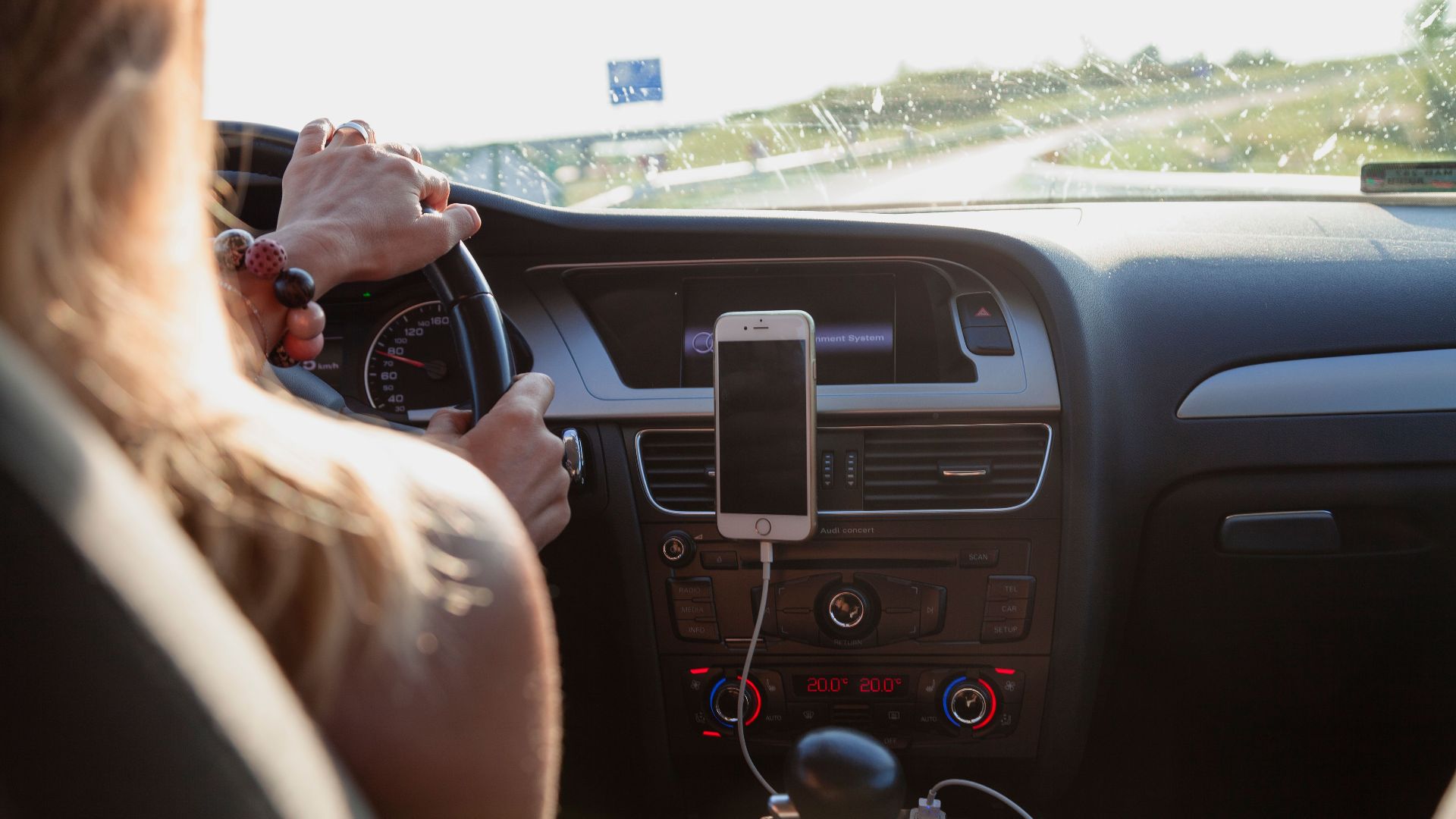 View from behind a woman driving a car on a sunny day with a phone mounted on the dashboard.