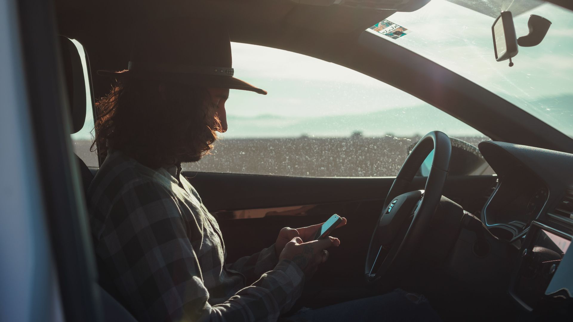 Profile of a man with a hat using a phone inside a car during the day.
