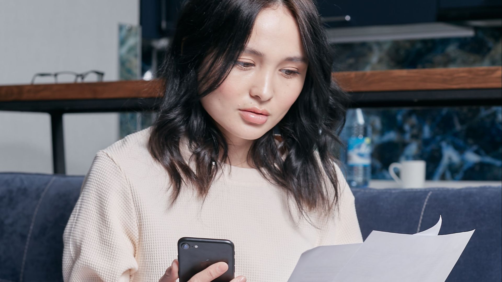 Young woman checking financial documents while sitting on a sofa with a smartphone and laptop.
