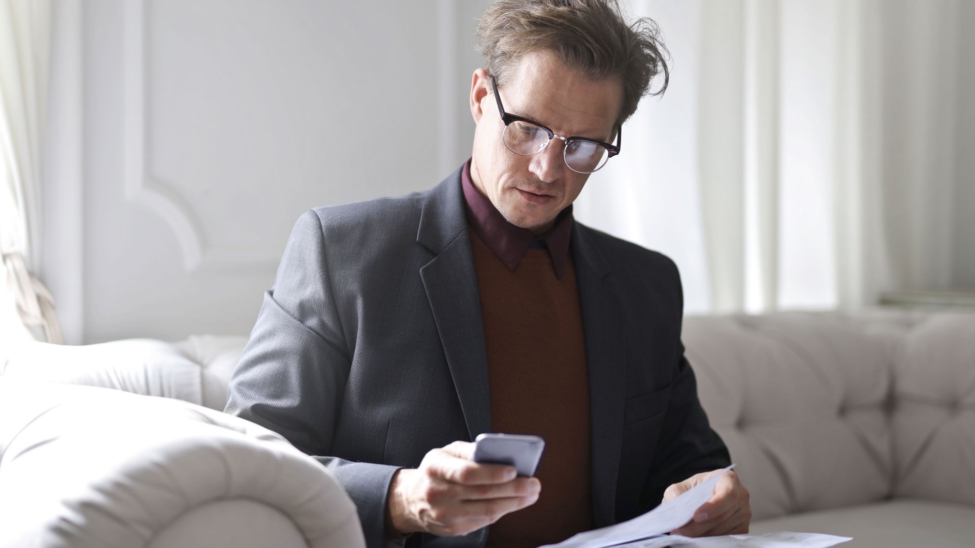 Focused businessman on a sofa using smartphone and reviewing documents.