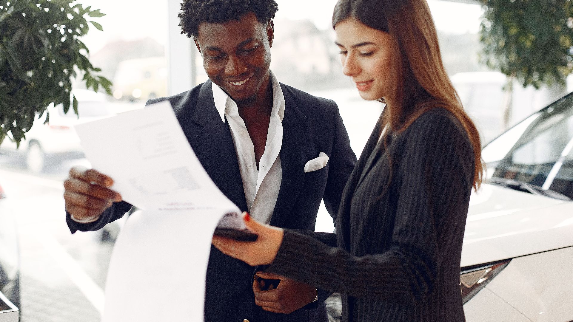 Cheerful smiling multiethnic businessman in classy suit and professional elegant female consultant standing close and reading contract details in car showroom