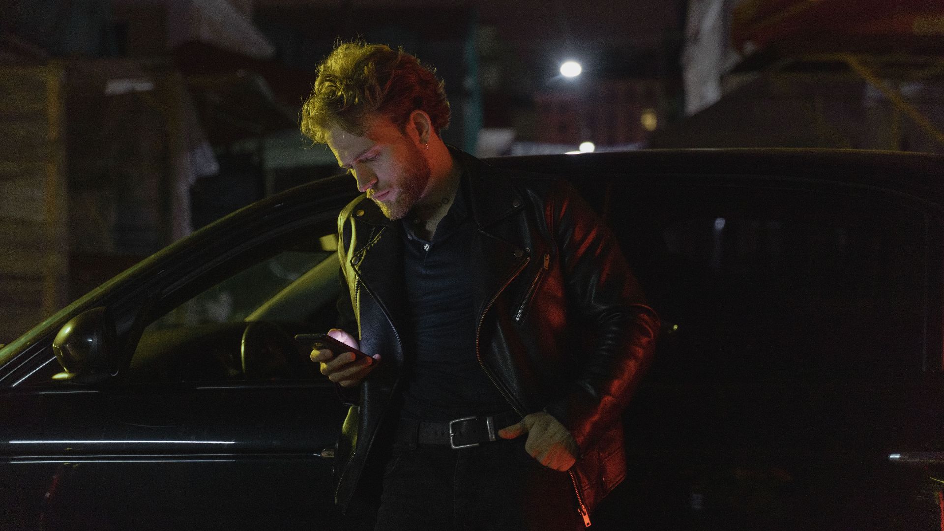 Caucasian man stands by a car at night, using his phone. Retro 80s style with a moody atmosphere.
