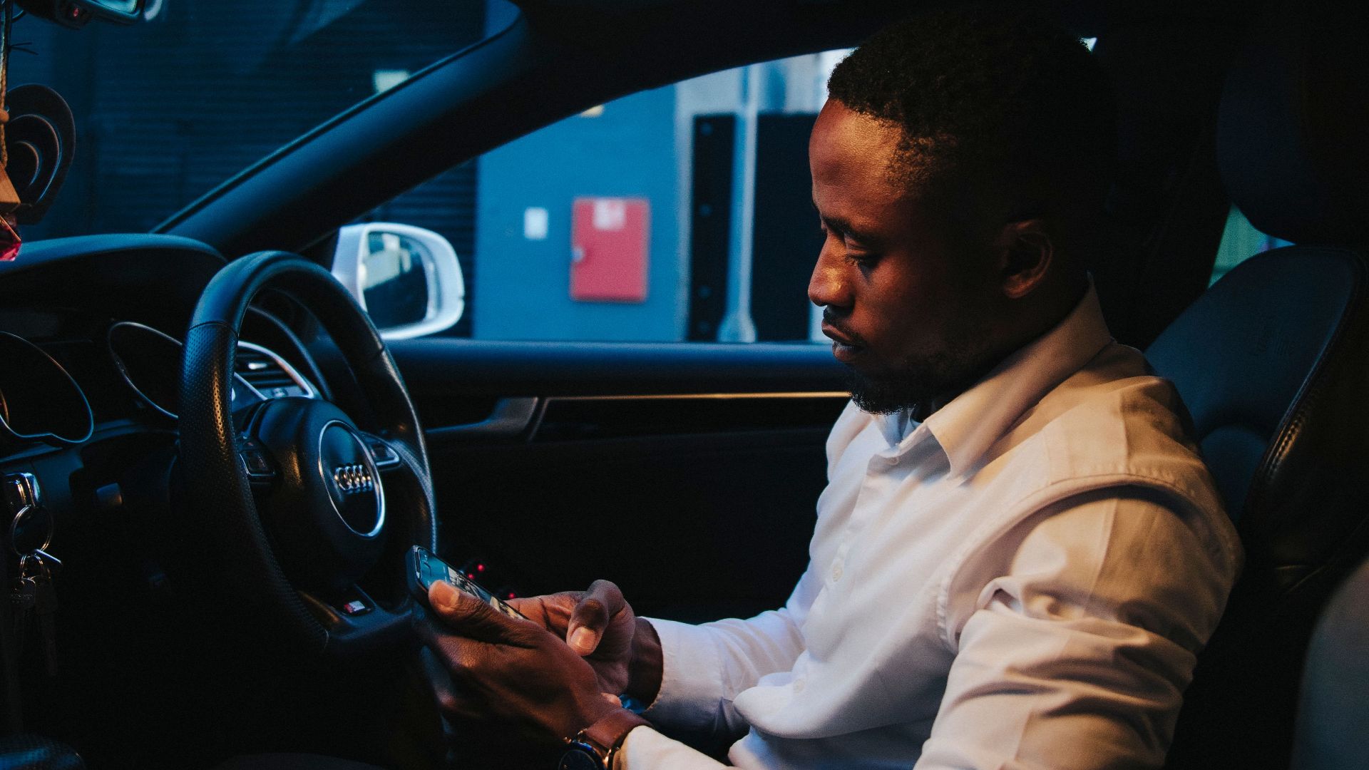 An adult man in a white shirt checking his smartphone while sitting in a car at night.