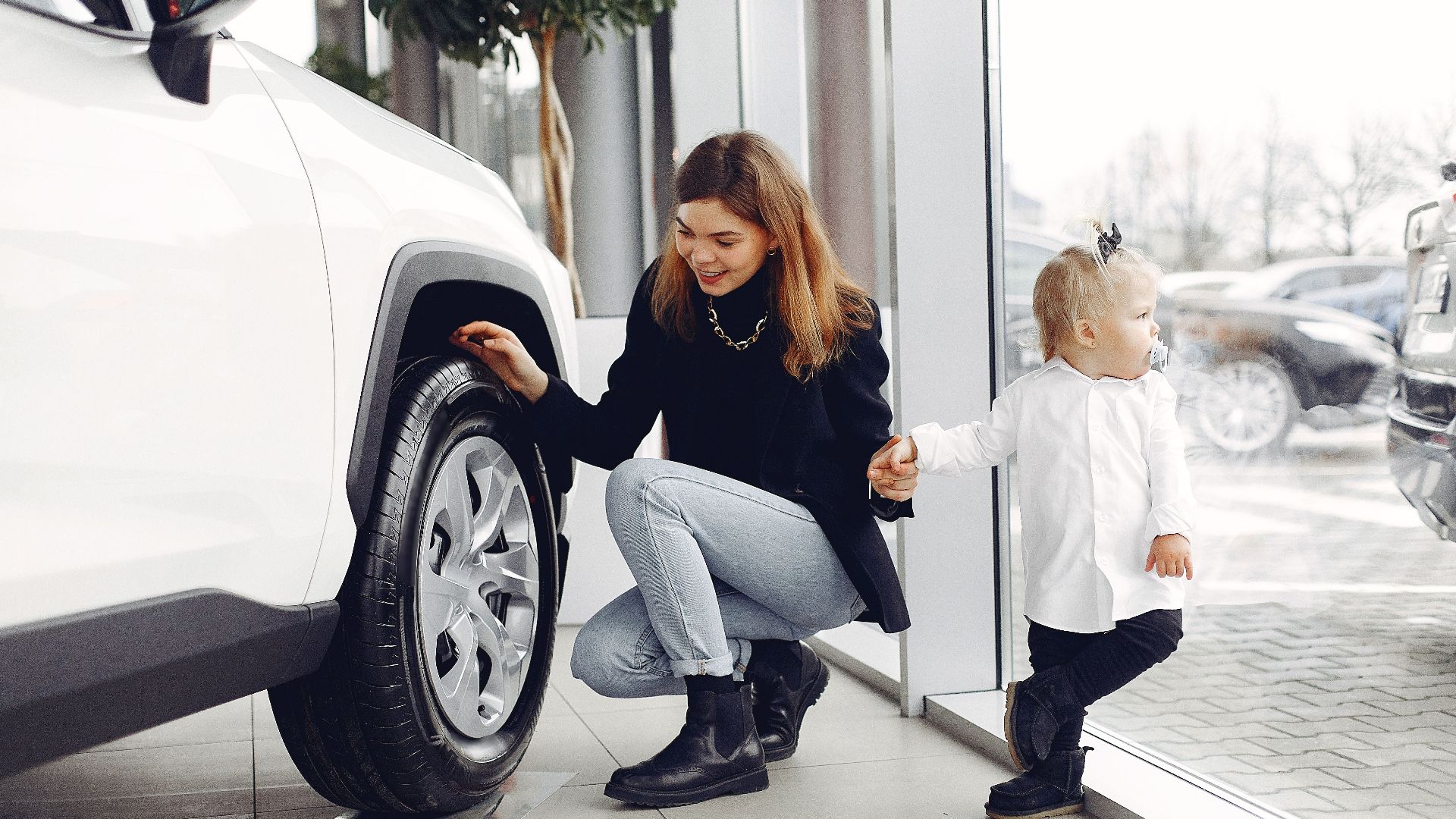 Full length of cheerful smiling mother in jeans holding hands with cute little girl and looking at car wheel in car showroom in daylight