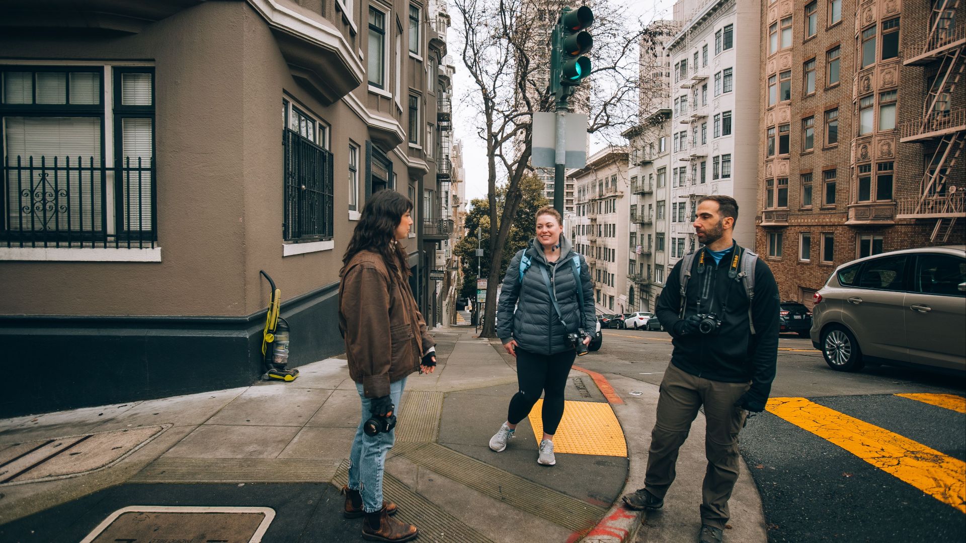 Three photographers with cameras chat on a city street corner amid urban architecture.