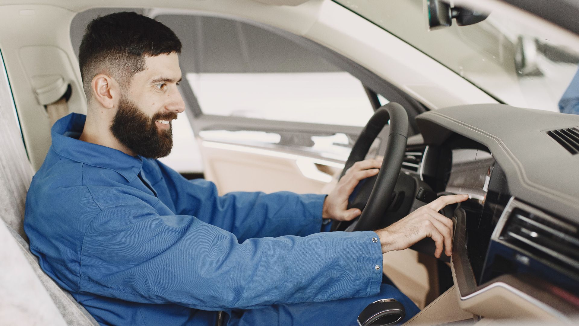 Mechanic in blue coveralls interacts with car dashboard, smiling and focused.