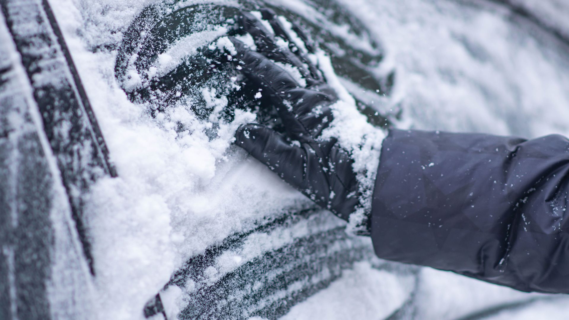 A close-up of a gloved hand clearing snow from a car window during winter.