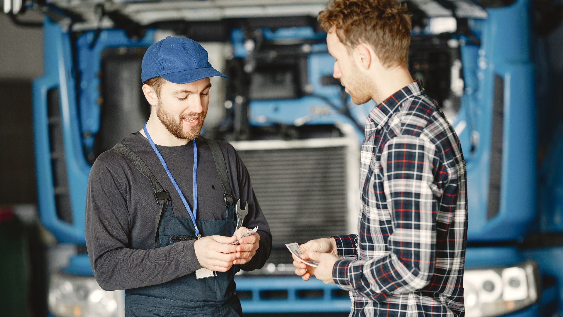Auto mechanic in blue uniform receives payment from customer in a garage setting with a truck in the background.
