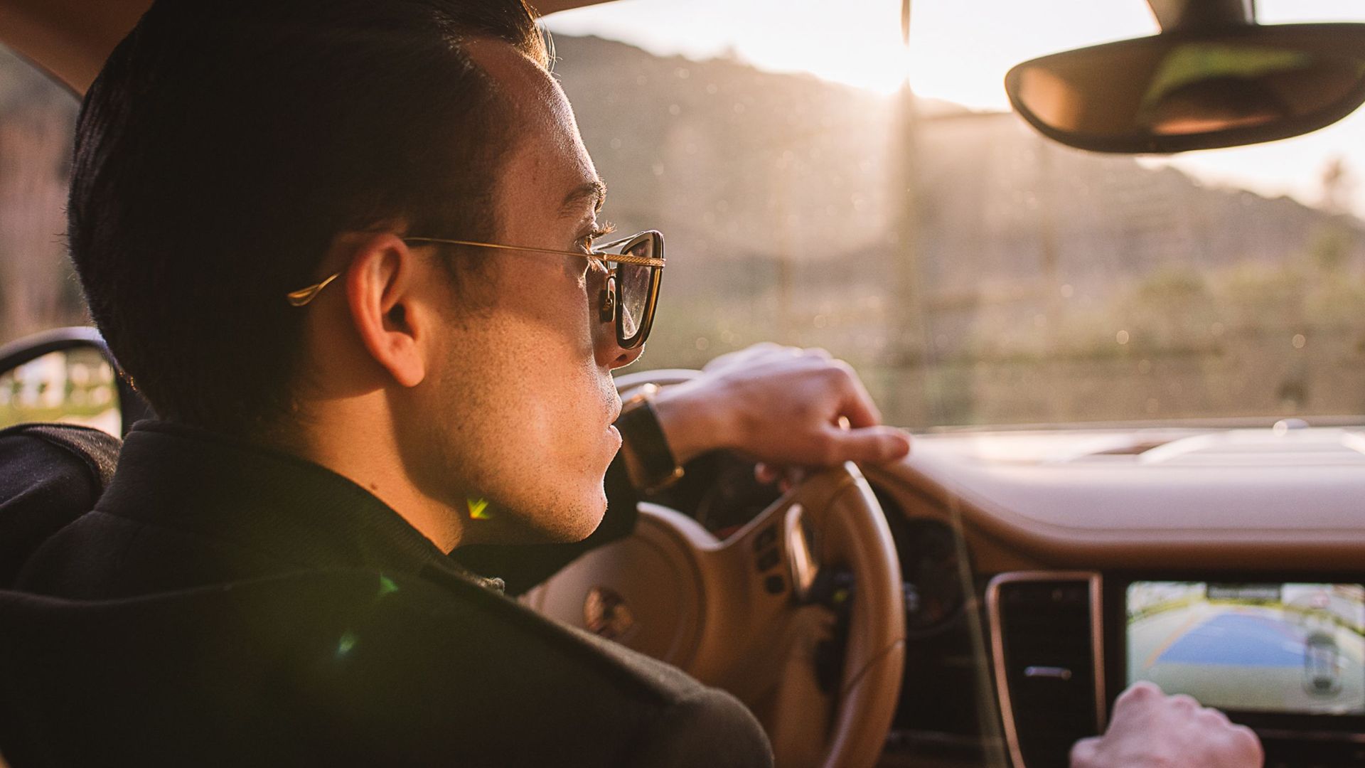 Man driving luxury car at sunset wearing sunglasses, showcasing elegant car interior and confident style.