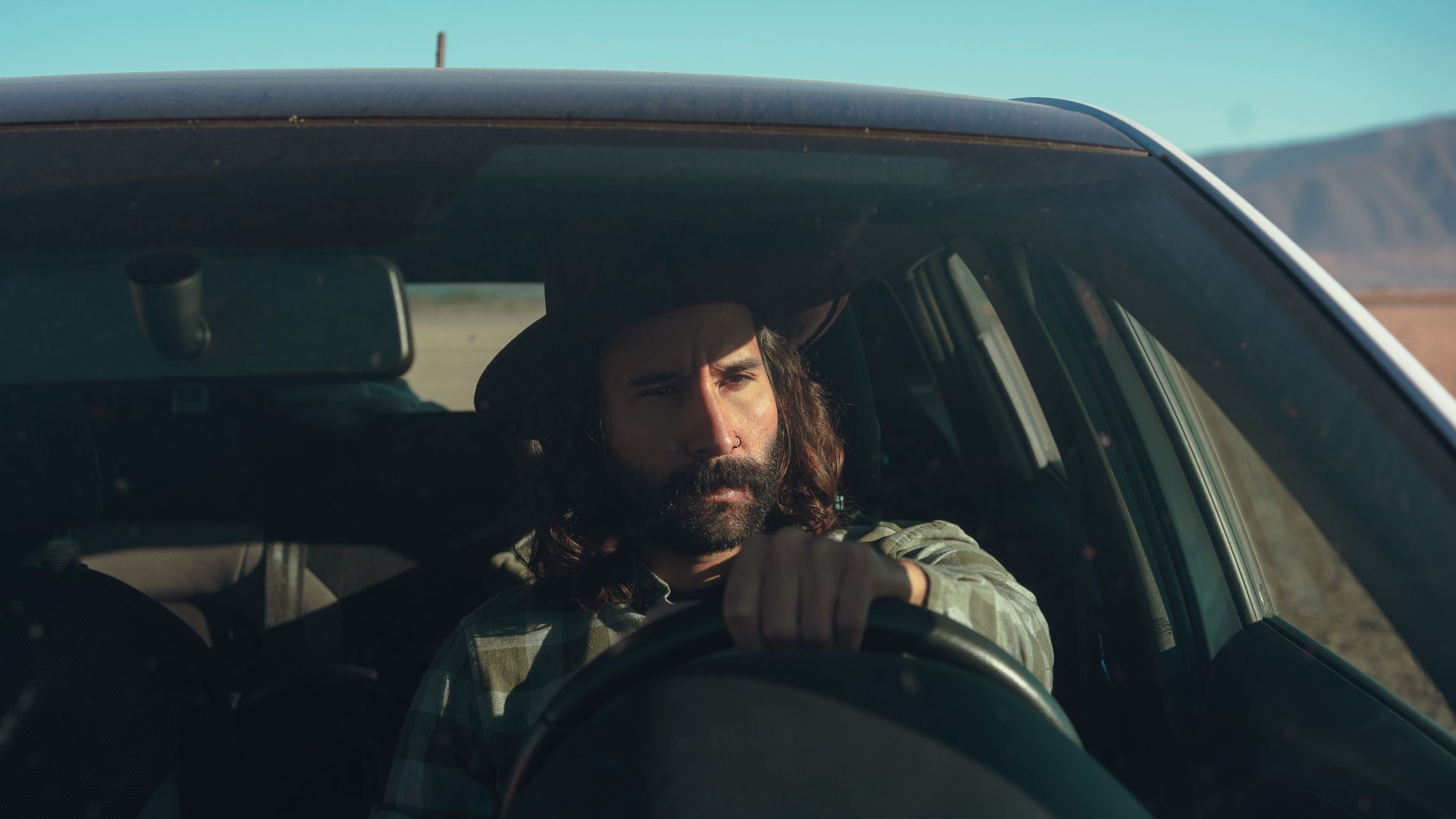 A bearded man in a hat driving a car through a sunny desert landscape, conveying a sense of adventure.