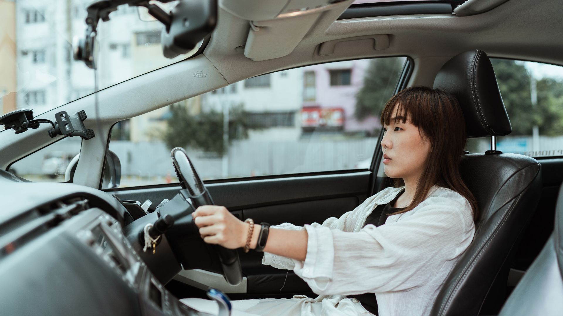 Side view unemotional Asian female in casual white clothes steering right hand drive car along urban streets in daytime