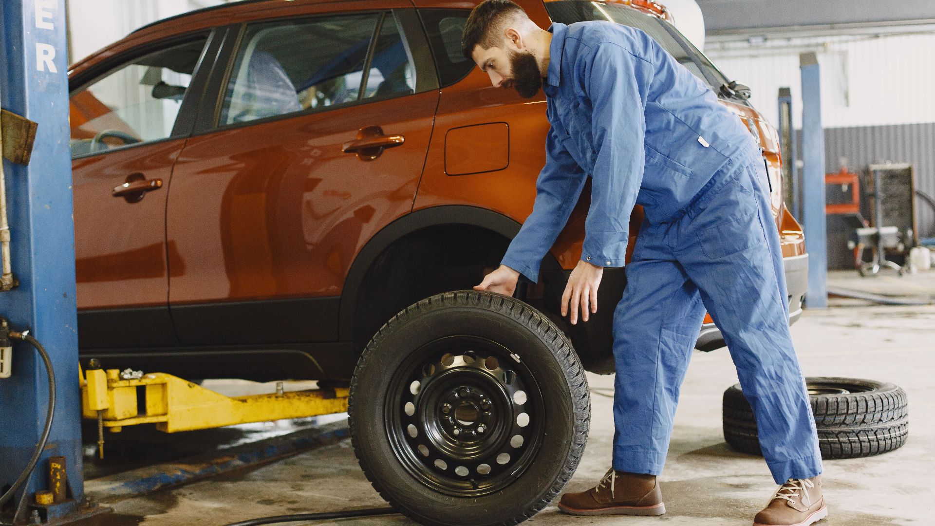 Mechanic in blue coveralls replacing tire on red SUV in repair shop garage.