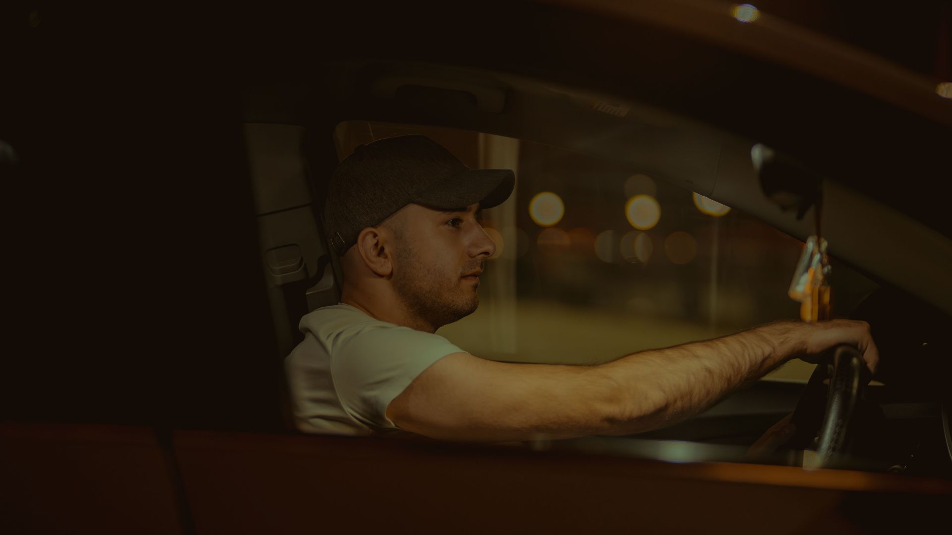 A man wearing a cap is driving a car at night, focused on the road ahead.