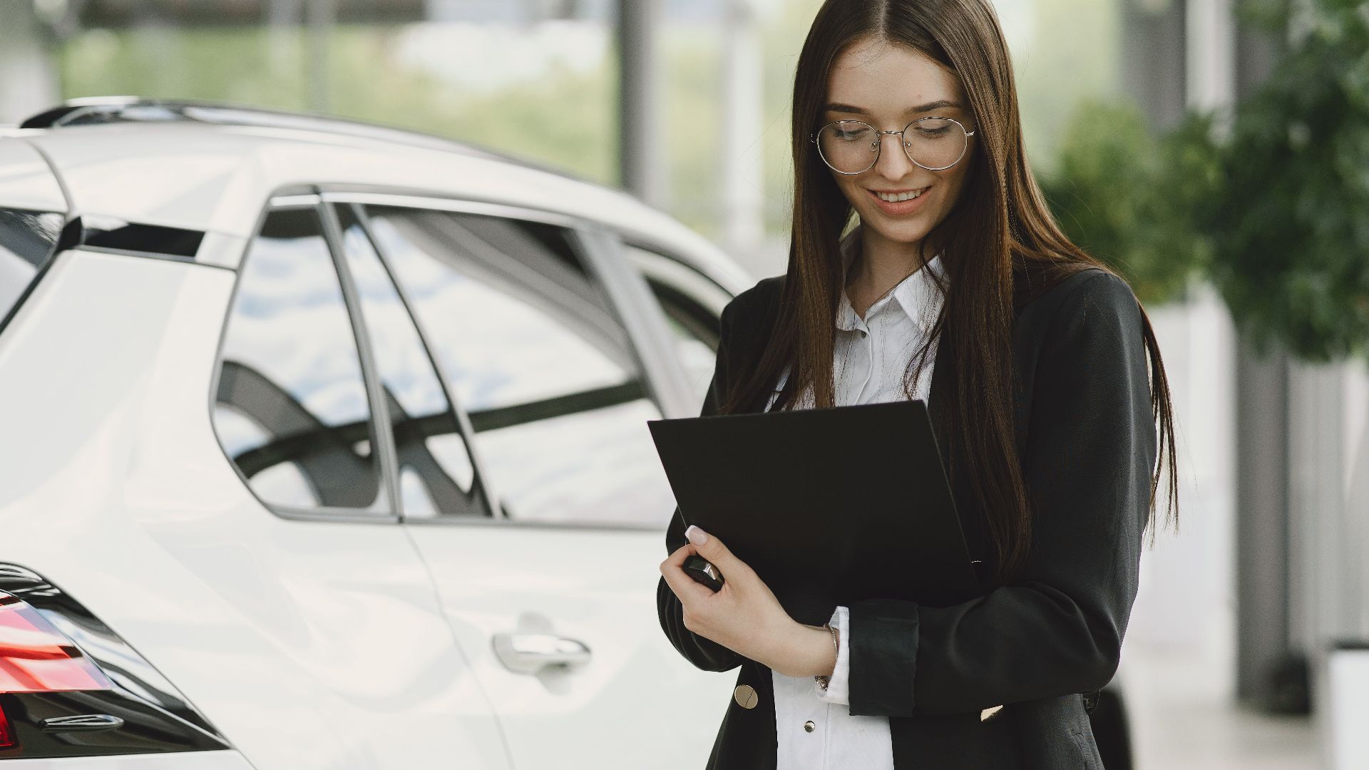 Smiling woman in a black jacket reviewing documents near a white car indoors.