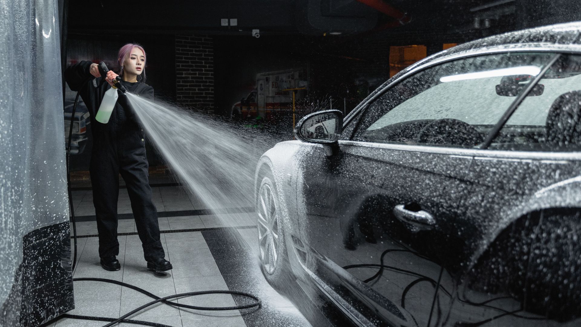 Woman using a pressure washer to clean a black car in an indoor car wash setting.