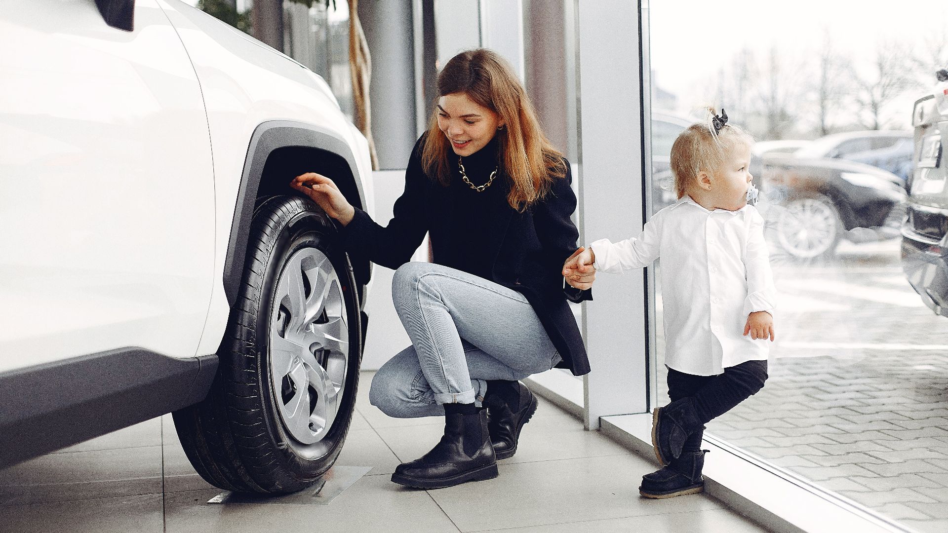 Full length of cheerful smiling mother in jeans holding hands with cute little girl and looking at car wheel in car showroom in daylight