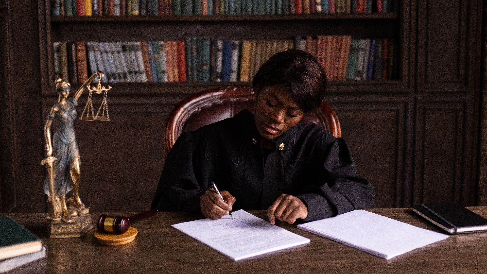 A judge in robes writing on a document at a desk in an office library with law books.