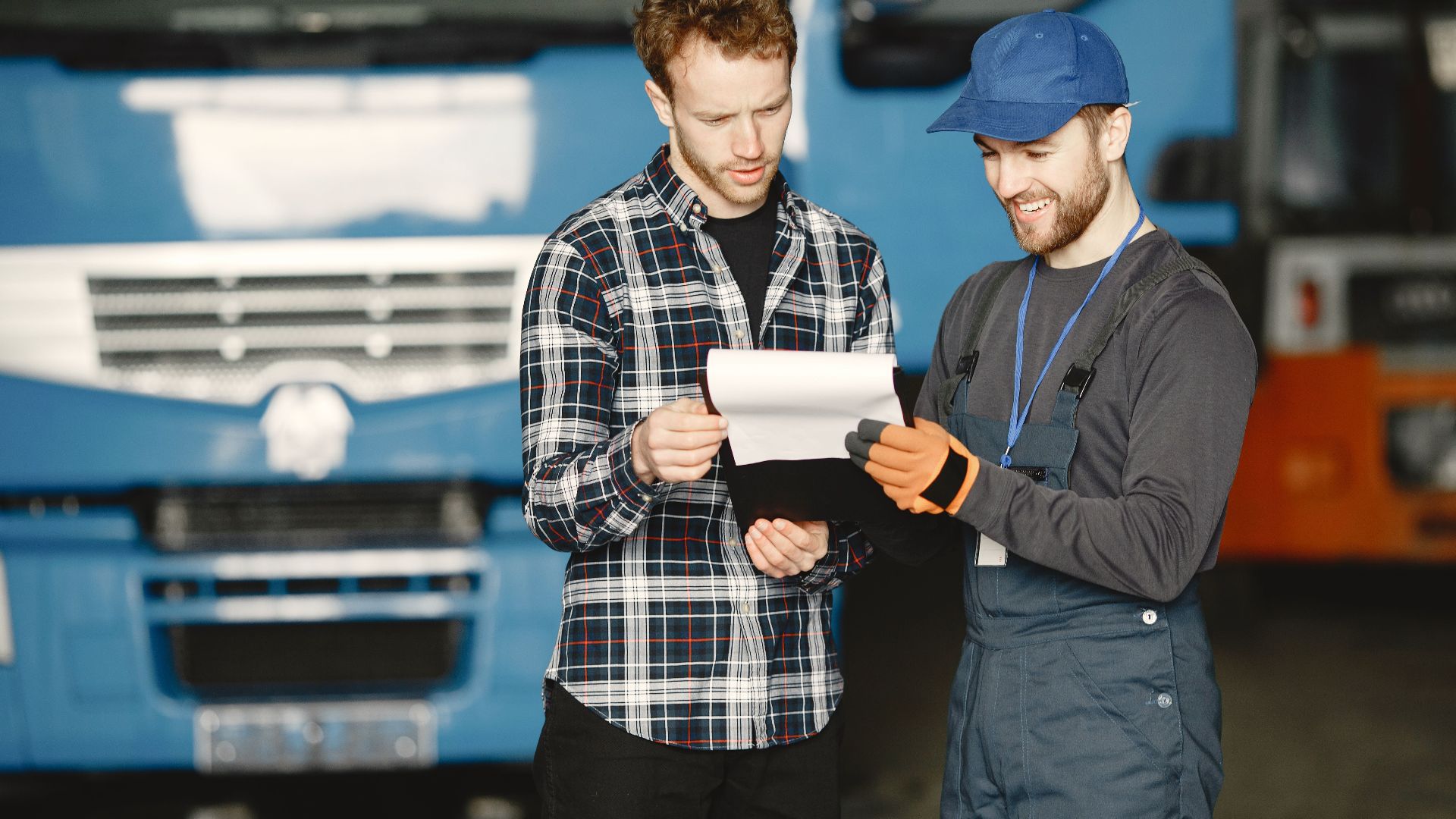 Two men in discussion over vehicle maintenance documents in a garage setting with trucks in the background.