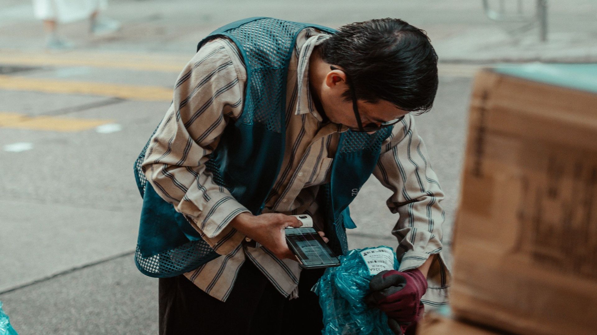 A street vendor examines packages on a busy city sidewalk, surrounded by boxes and pedestrians.