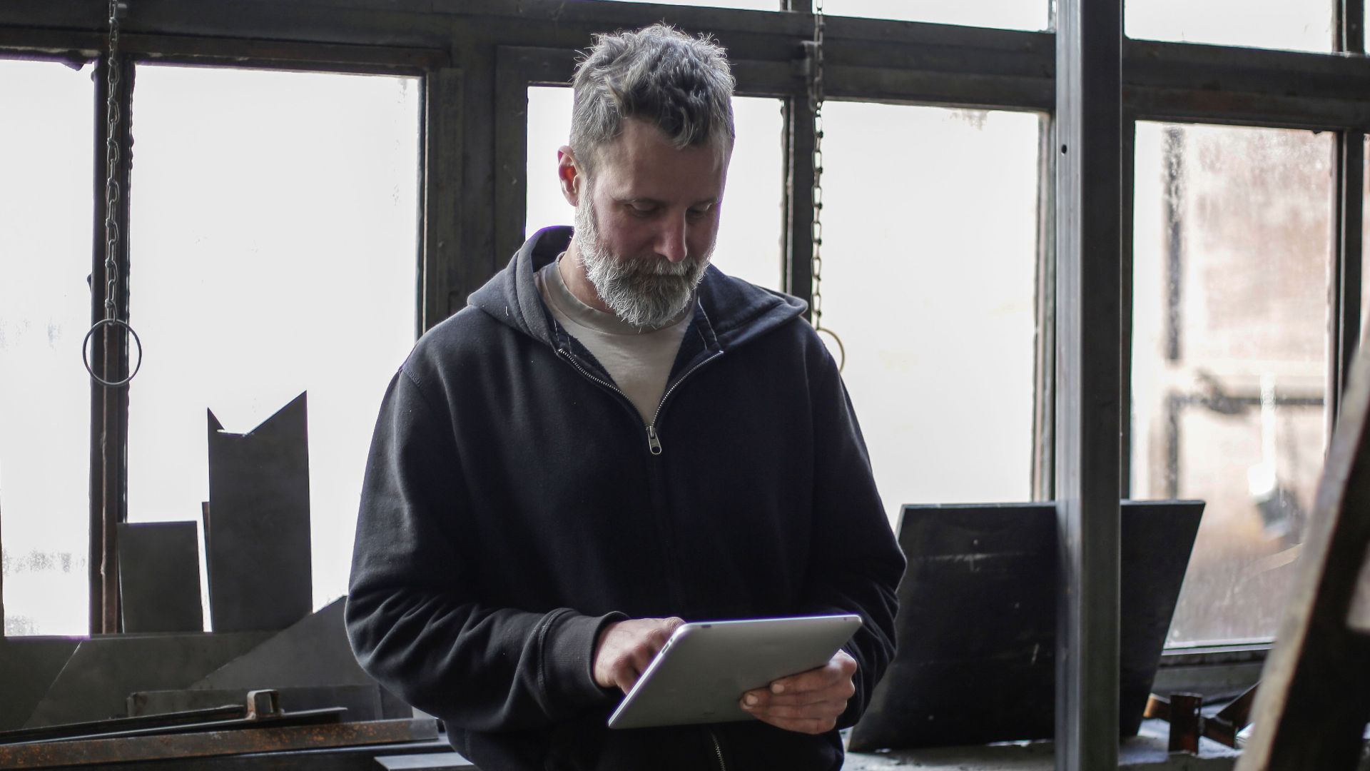 Serious adult bearded workman wearing workwear standing near window and browsing tablet while working in workshop