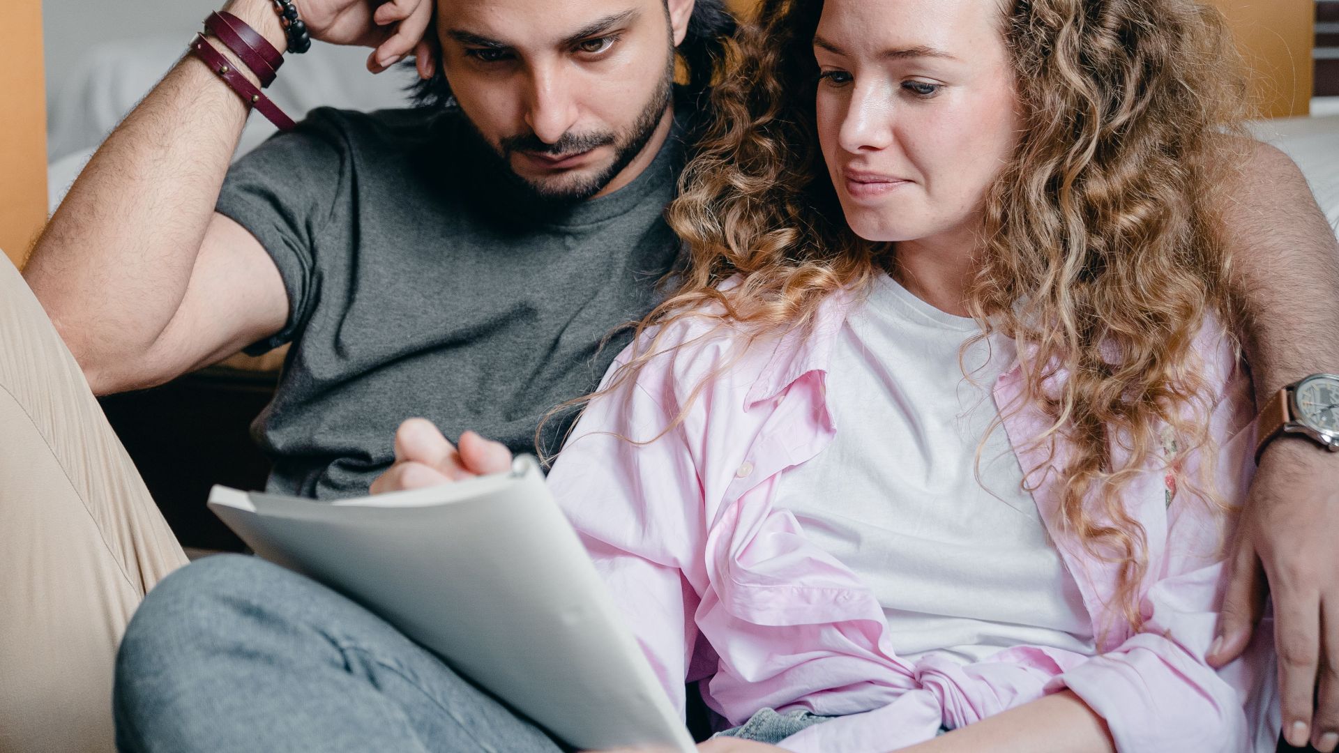 Pensive male and female in casual clothes sitting together among boxes and writing notes in notebook while leaning on bed
