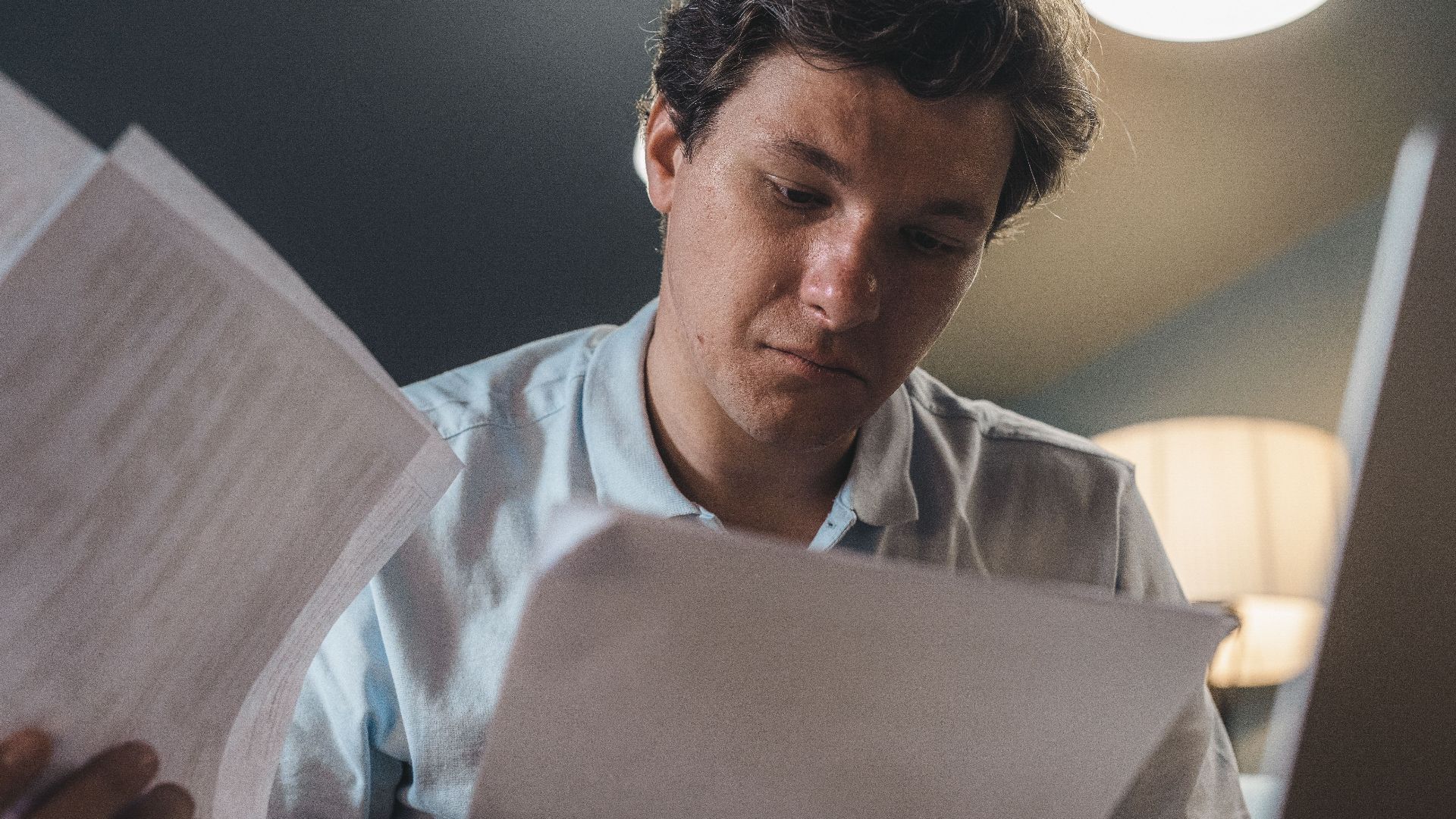 Serious man wearing a light blue shirt intently reading printed papers indoors under warm lighting.