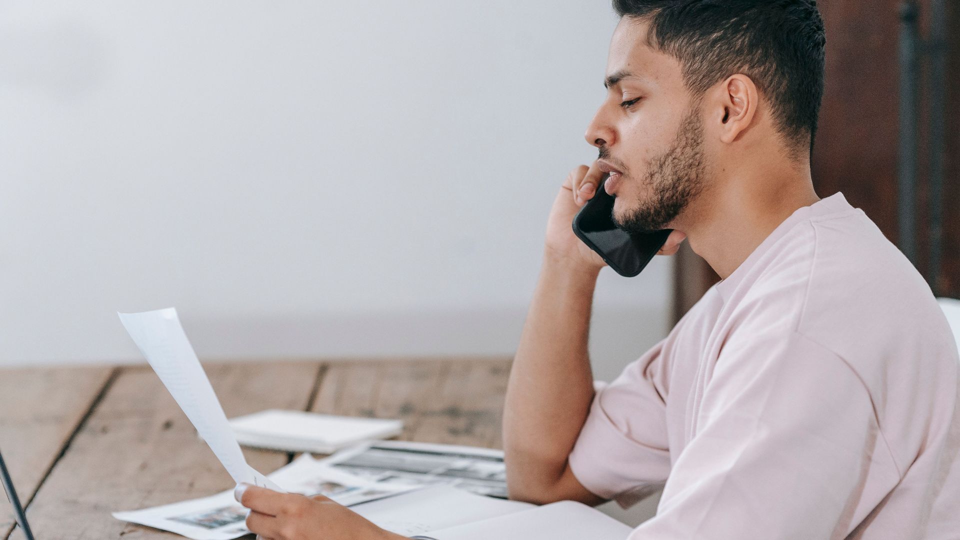 Side view of young Hispanic male remote employee discussing documents during phone call while working online with laptop and papers in home office