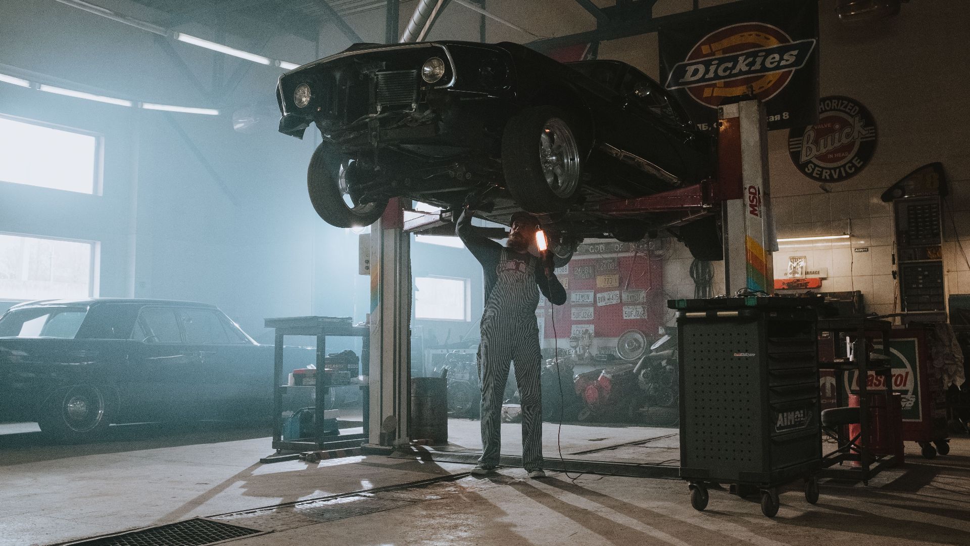 Male mechanic using a light to inspect a classic car lifted in an automotive garage.