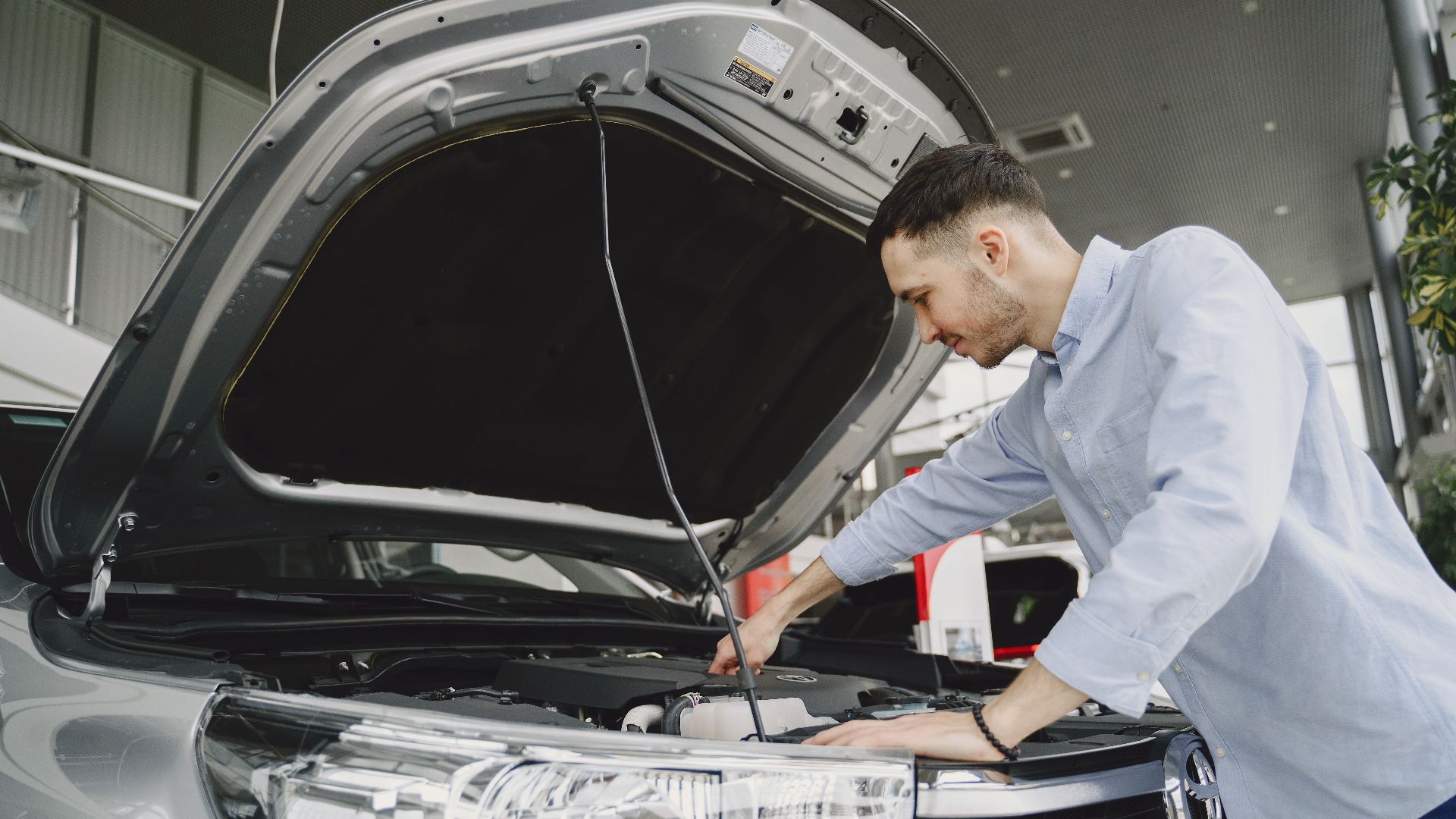 A man in a blue shirt examines a car's engine in a modern vehicle showroom.