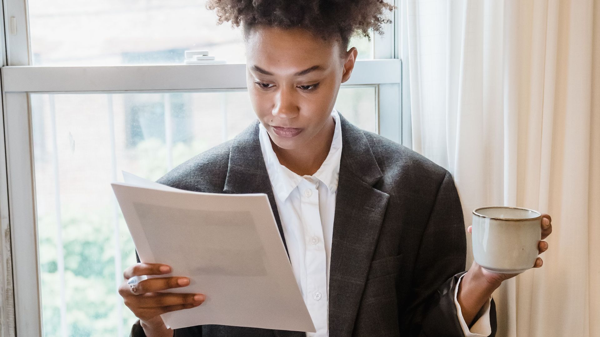 A focused woman reads a document while holding a drink by a sunlit window indoors.