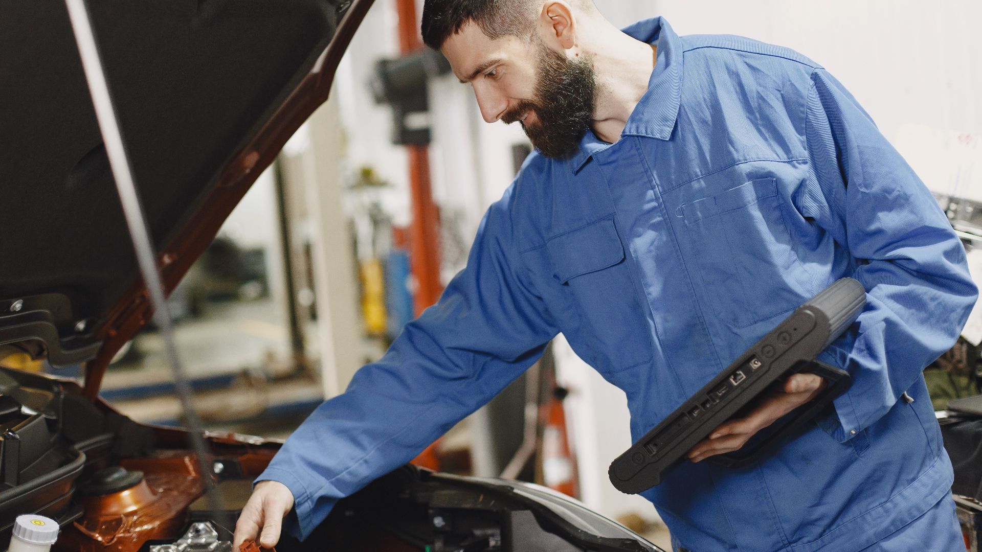 An auto mechanic in blue coveralls inspecting a car's engine in a garage.