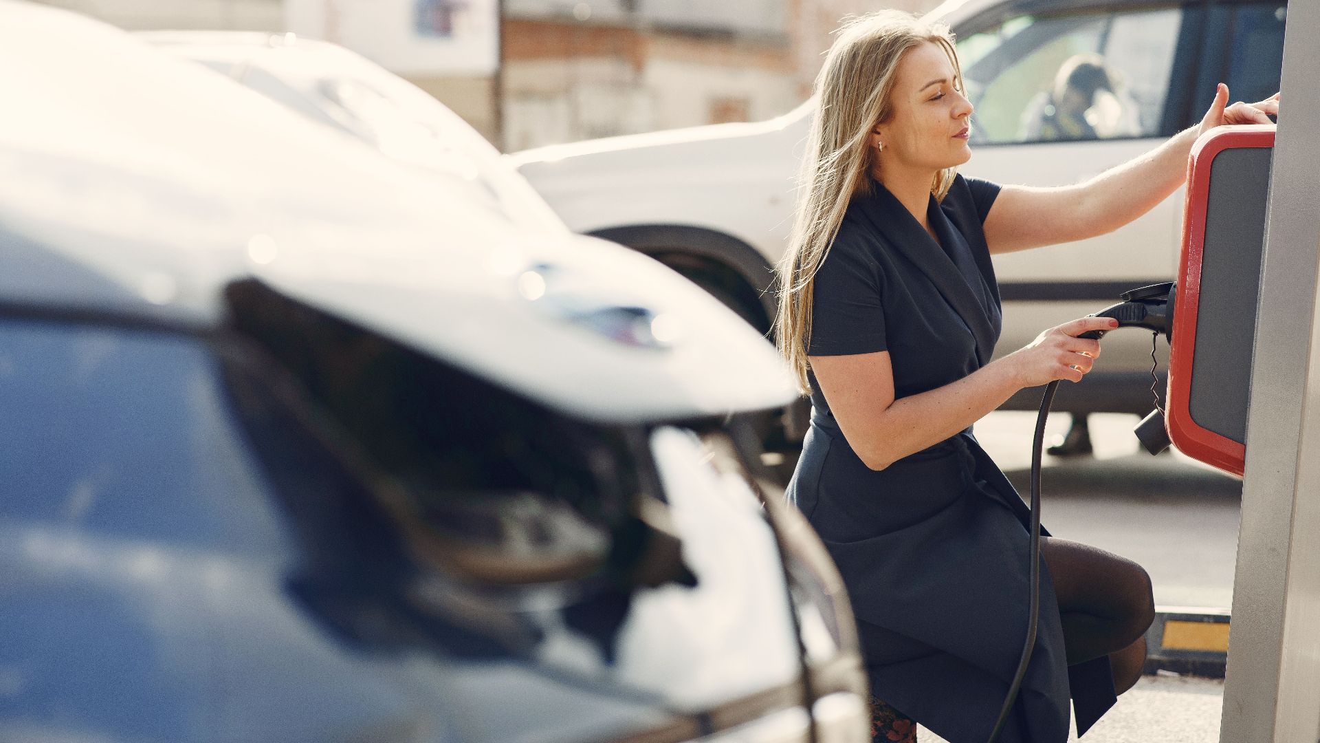 Side view of full length female driver in casual dress squatting near station while charging electric automobile for continuation of driving