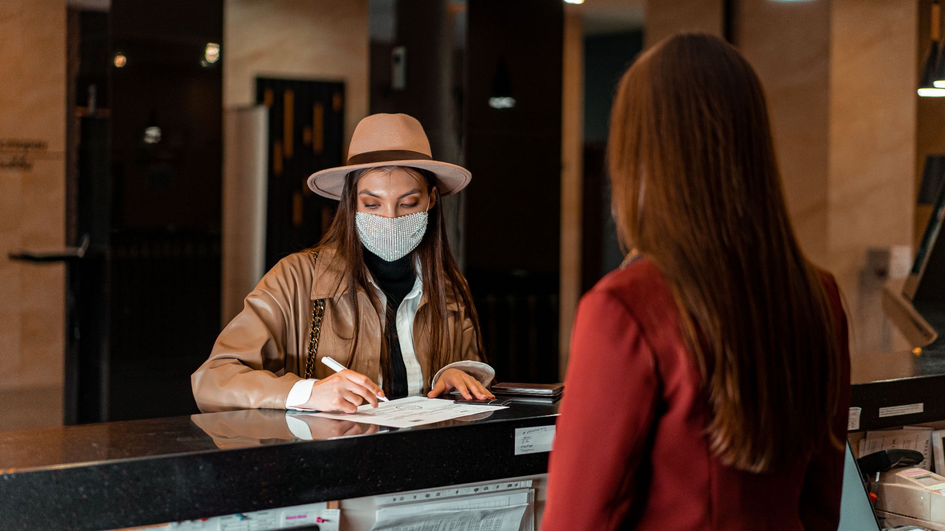 A woman in a hat and face mask checks in at a hotel reception, interacting with a receptionist.