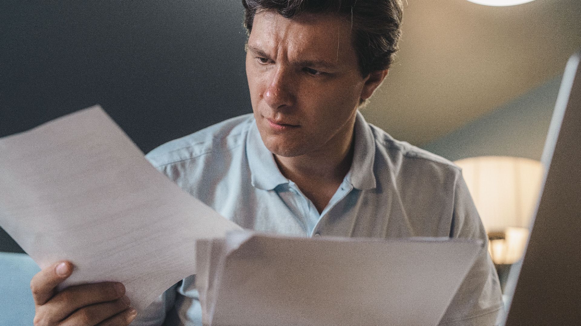 A man intently examines papers, seated indoors under warm lighting, focusing on his work.