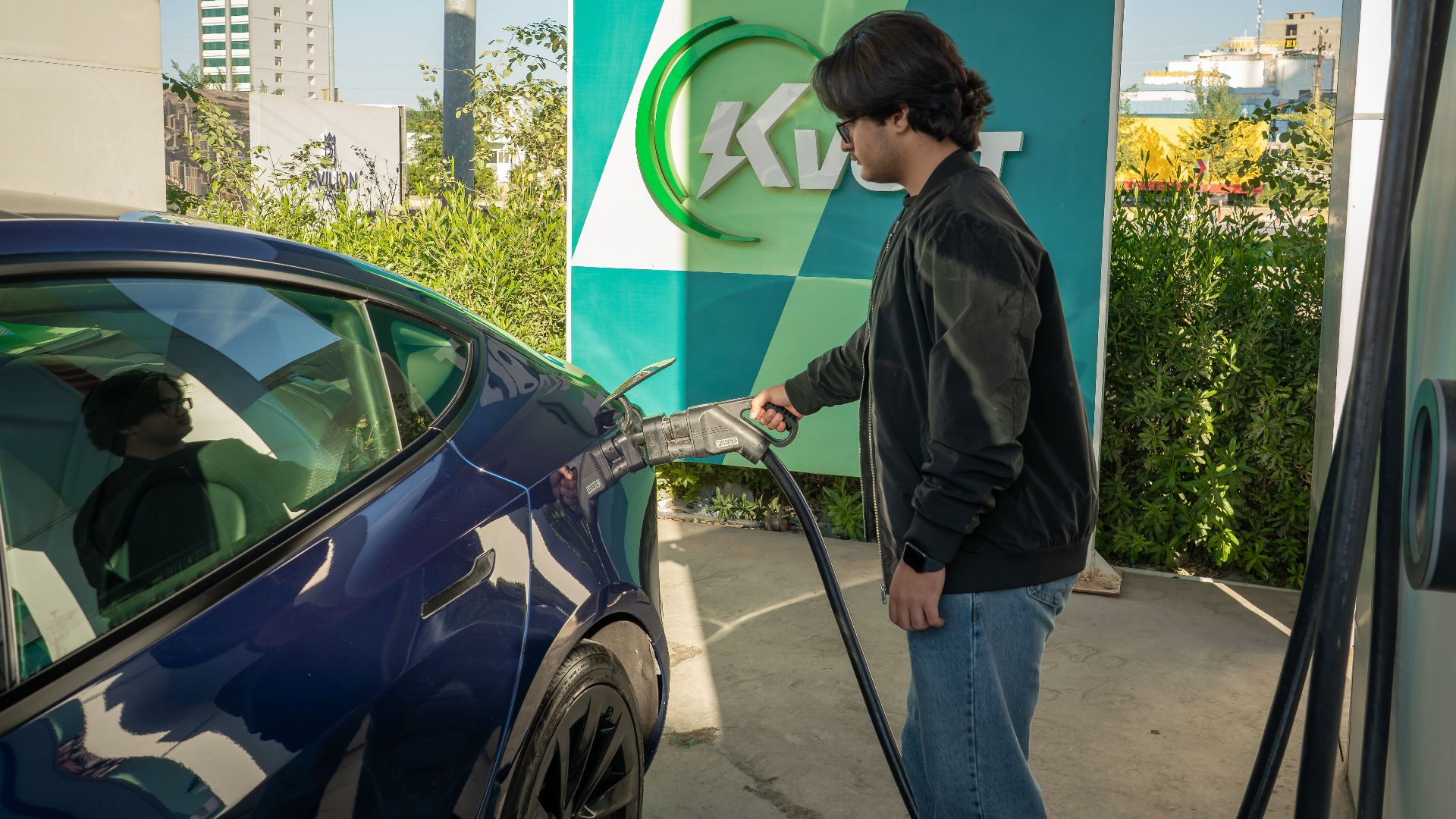 Young man charging a Tesla Model 3 at an outdoor electric vehicle station during the day.