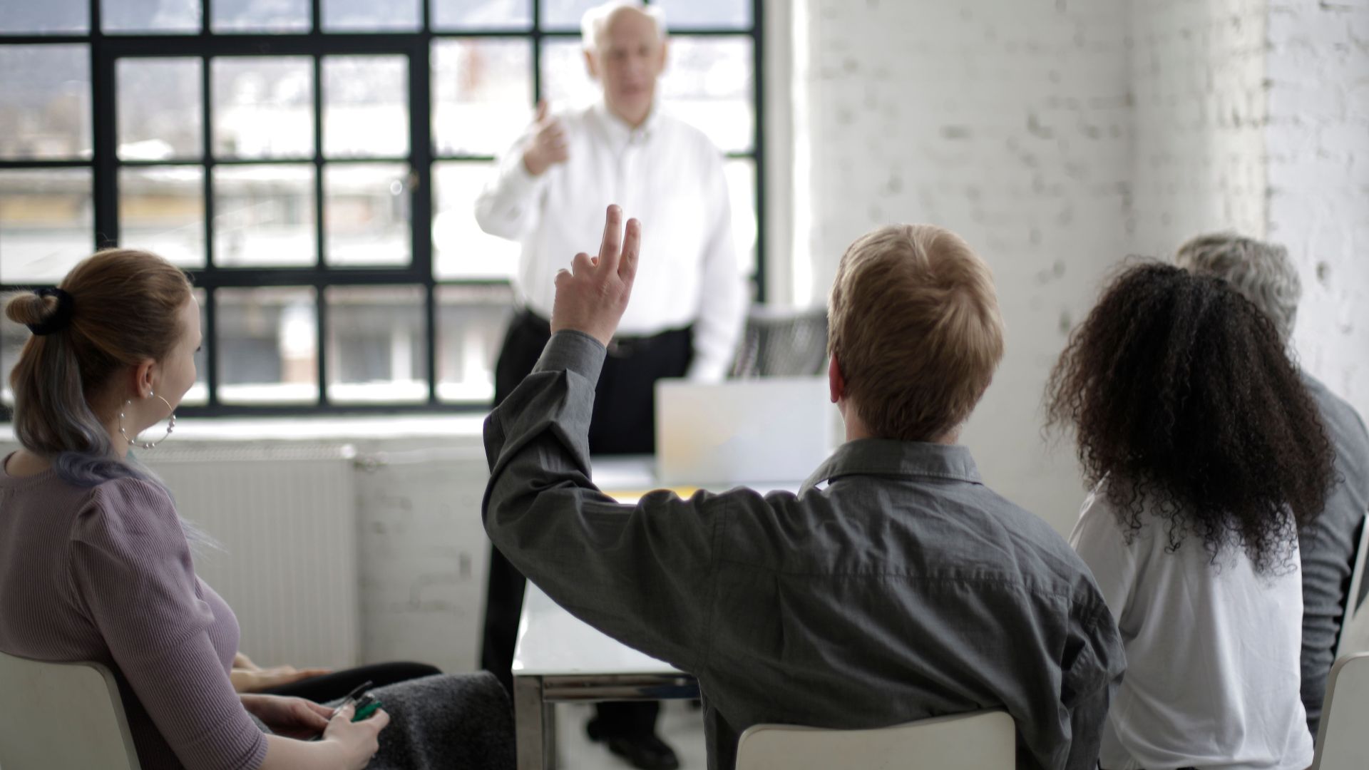Group of adults in a discussion, with one person raising a hand in a bright office space.