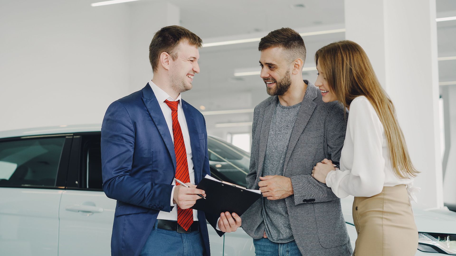A smiling couple buys a new car from a confident salesman inside a modern car dealership.