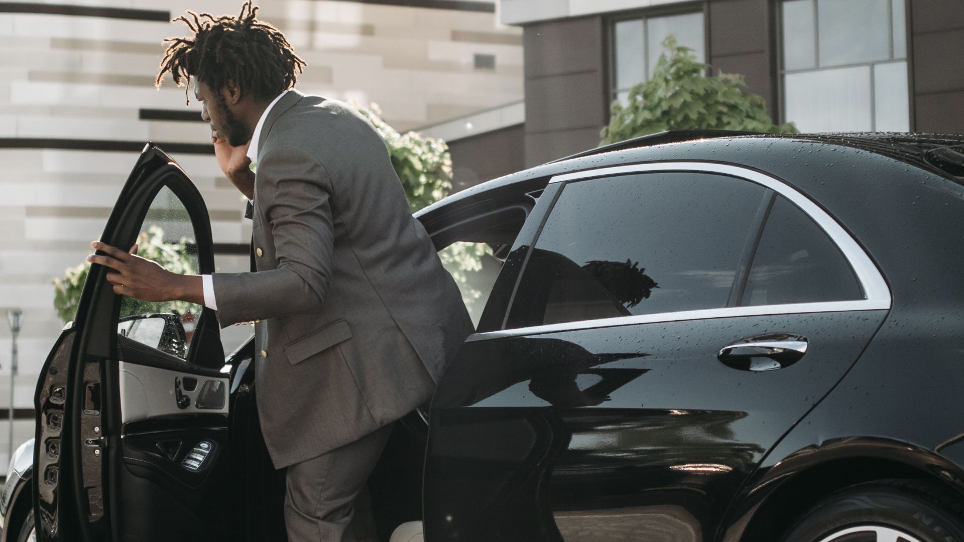 Stylish adult man in gray suit stepping out of a luxury black car in a modern city environment.