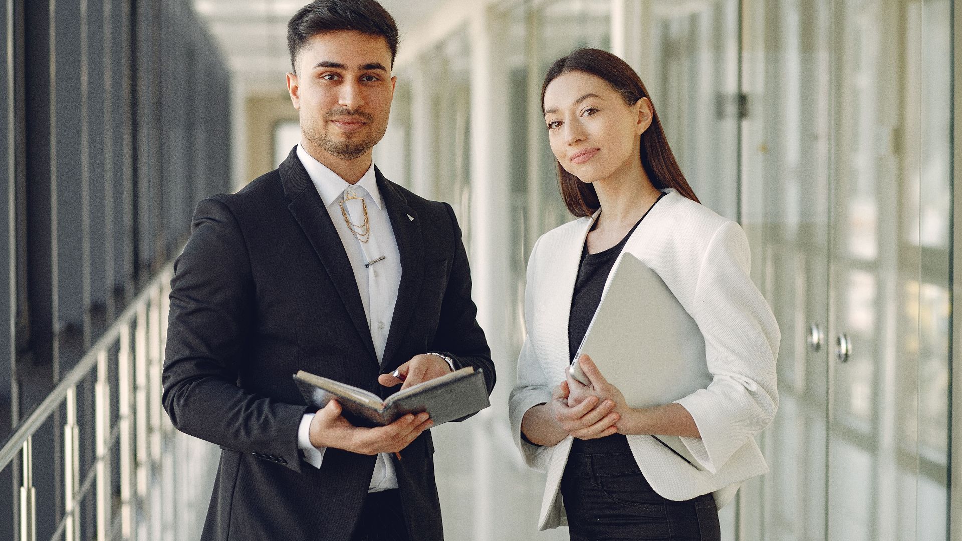 Confident well dressed multiracial coworkers standing in modern office corridor while holding laptop and notebook in hands and looking at camera