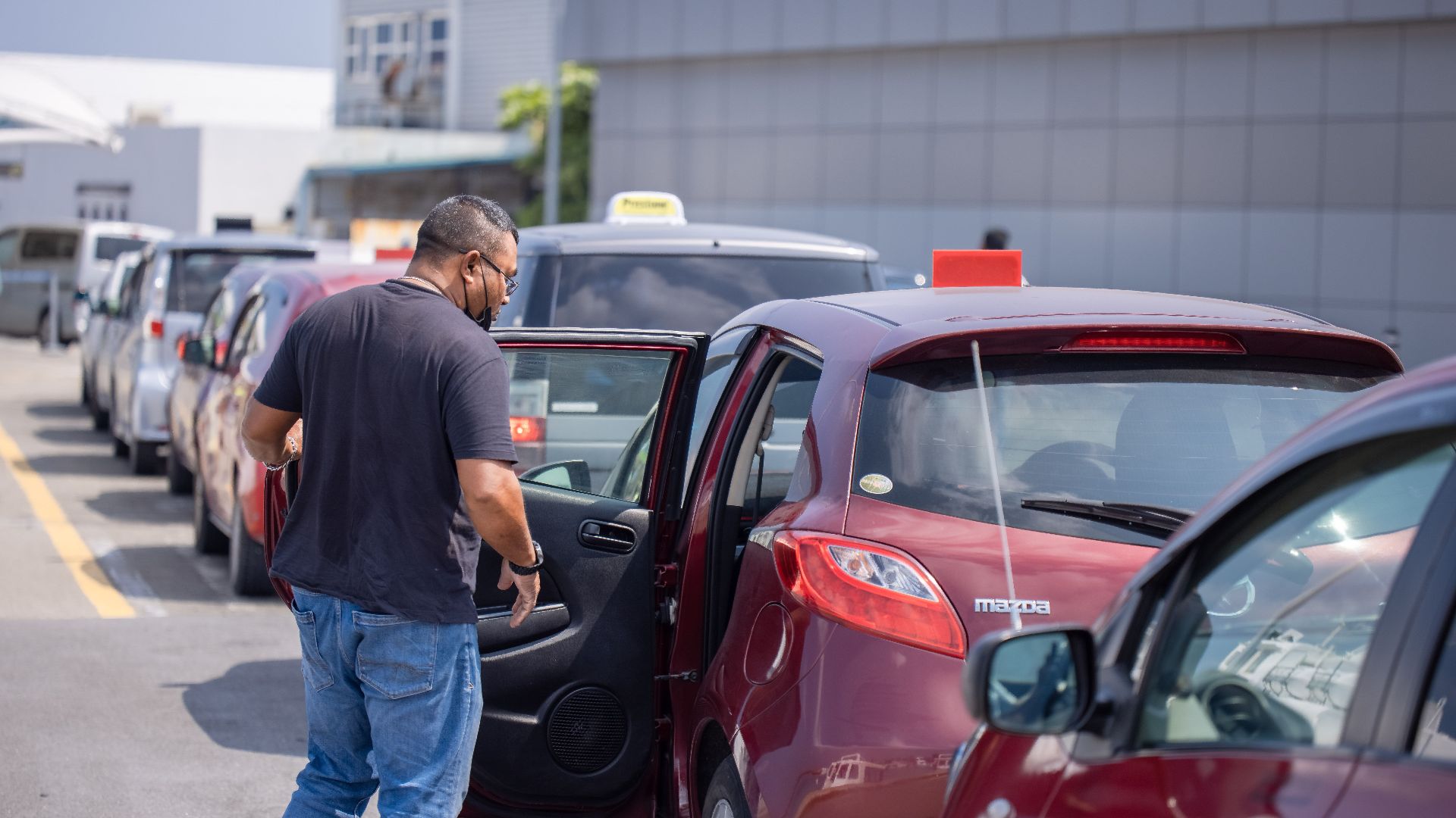 A man enters a red Mazda taxi in a line of parked cars on a sunny day.