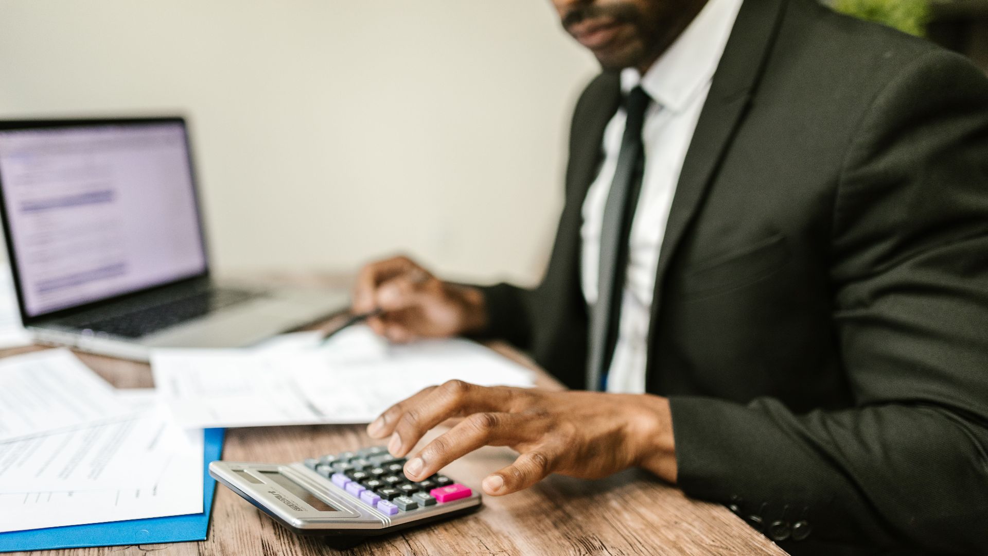 A businessman in a black suit using a calculator at a desk with financial documents and a laptop.
