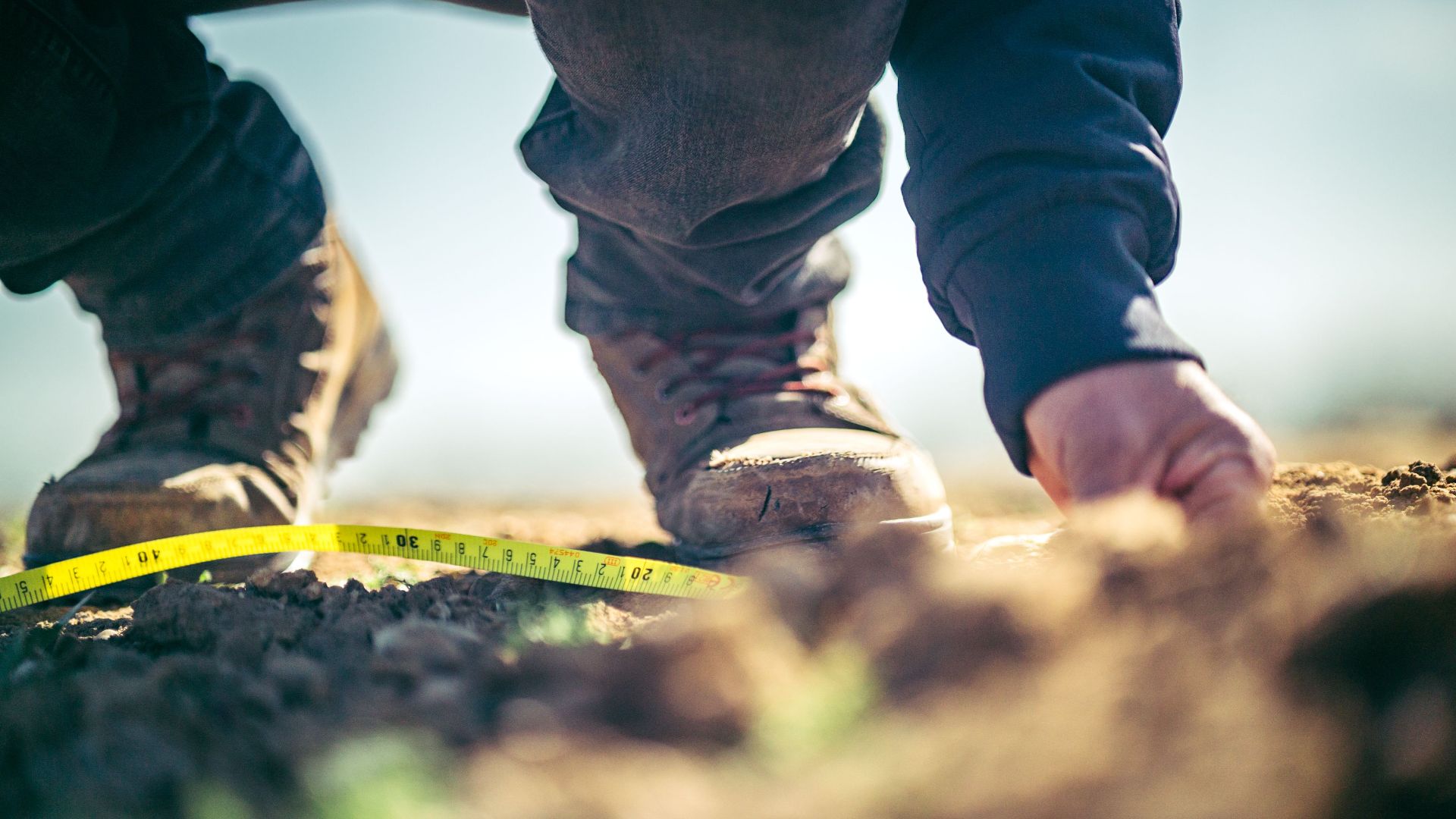 Focused shot of a person in boots measuring soil with tape outdoors.
