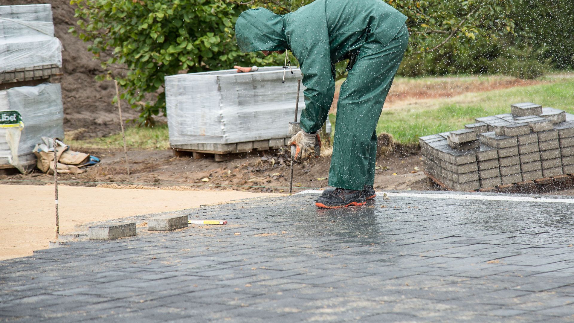 A worker in protective gear lays paving stones on a wet day.