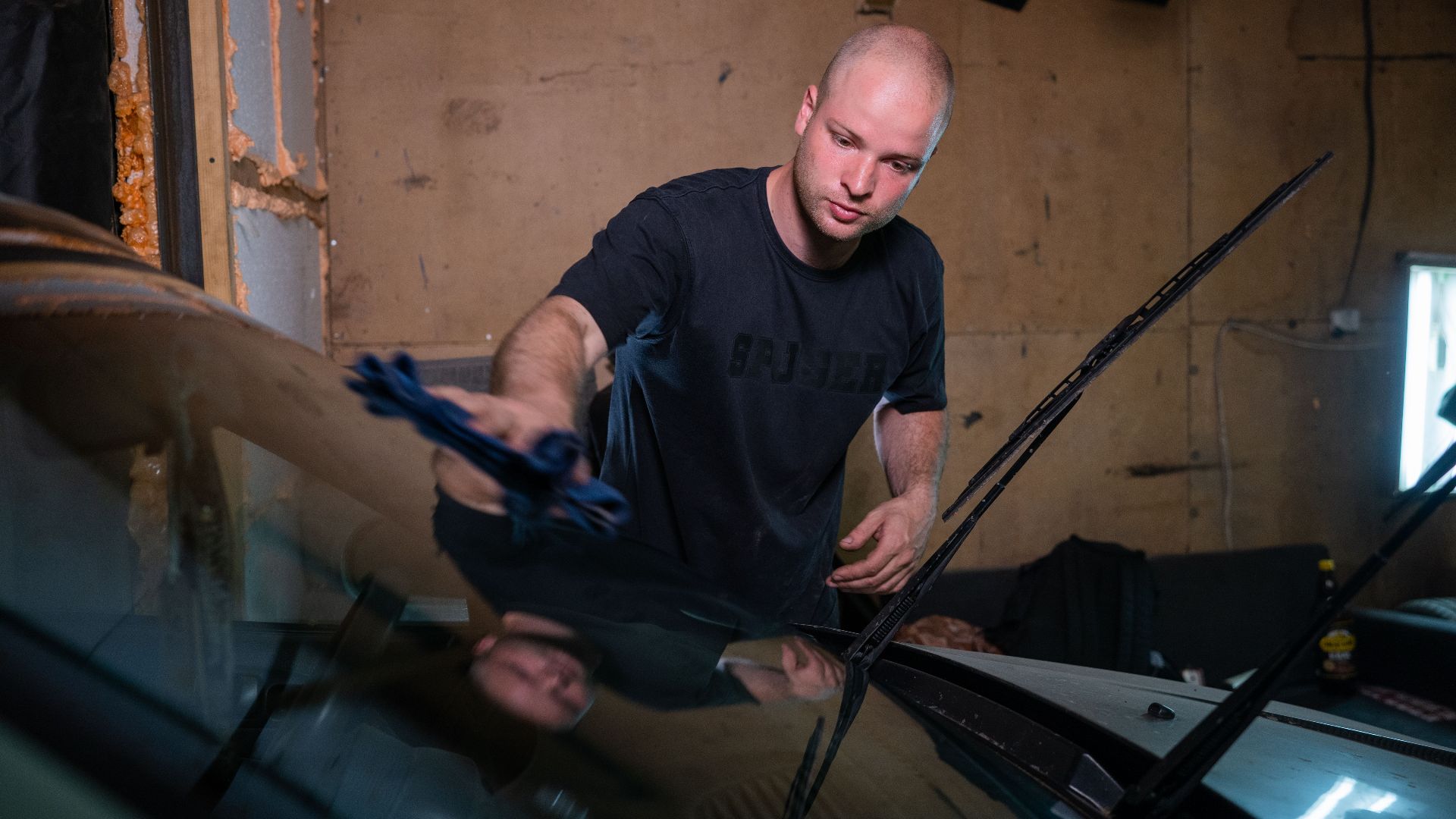 A man wipes a car windshield inside a garage, focused on cleaning.