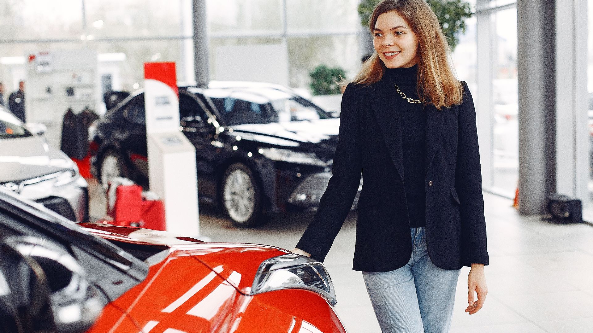Happy stylish young female in jeans and black jacket standing near modern red shiny car in contemporary car showroom and looking at purchase dreamily